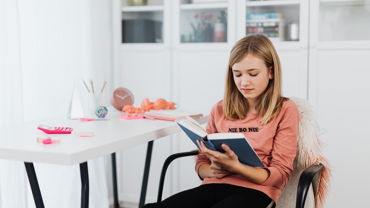 Teen girl in pink shirt reads book in bright, modern room for study or relaxation.